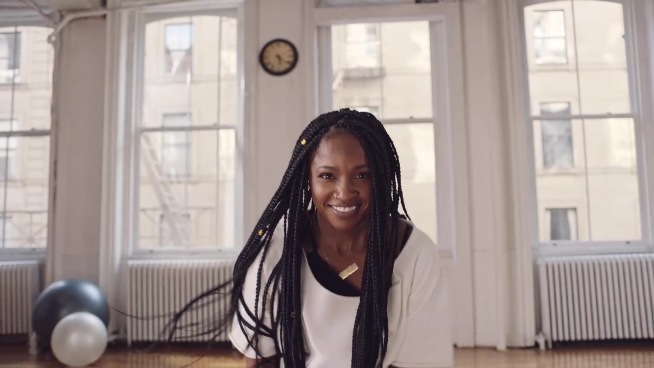 Smiling person with long braids leaning toward the camera in a bright window-lined studio