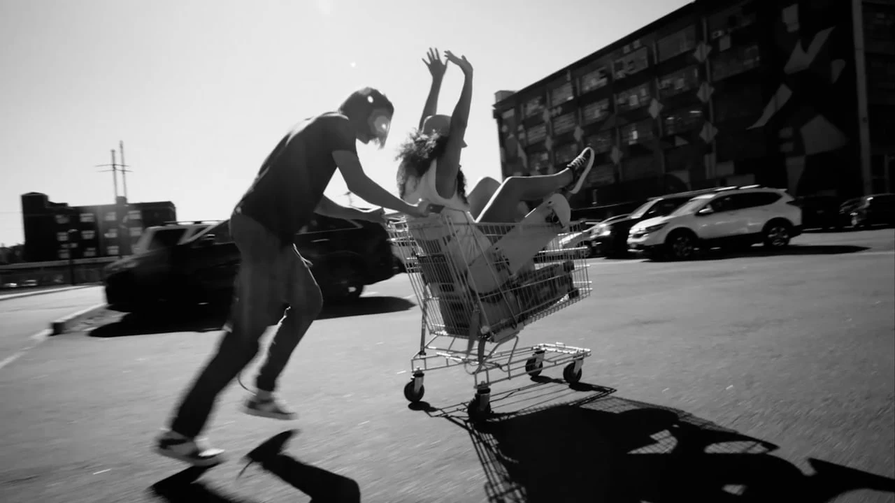 Black-and-white scene of two people racing through a parking lot with one riding in a shopping cart