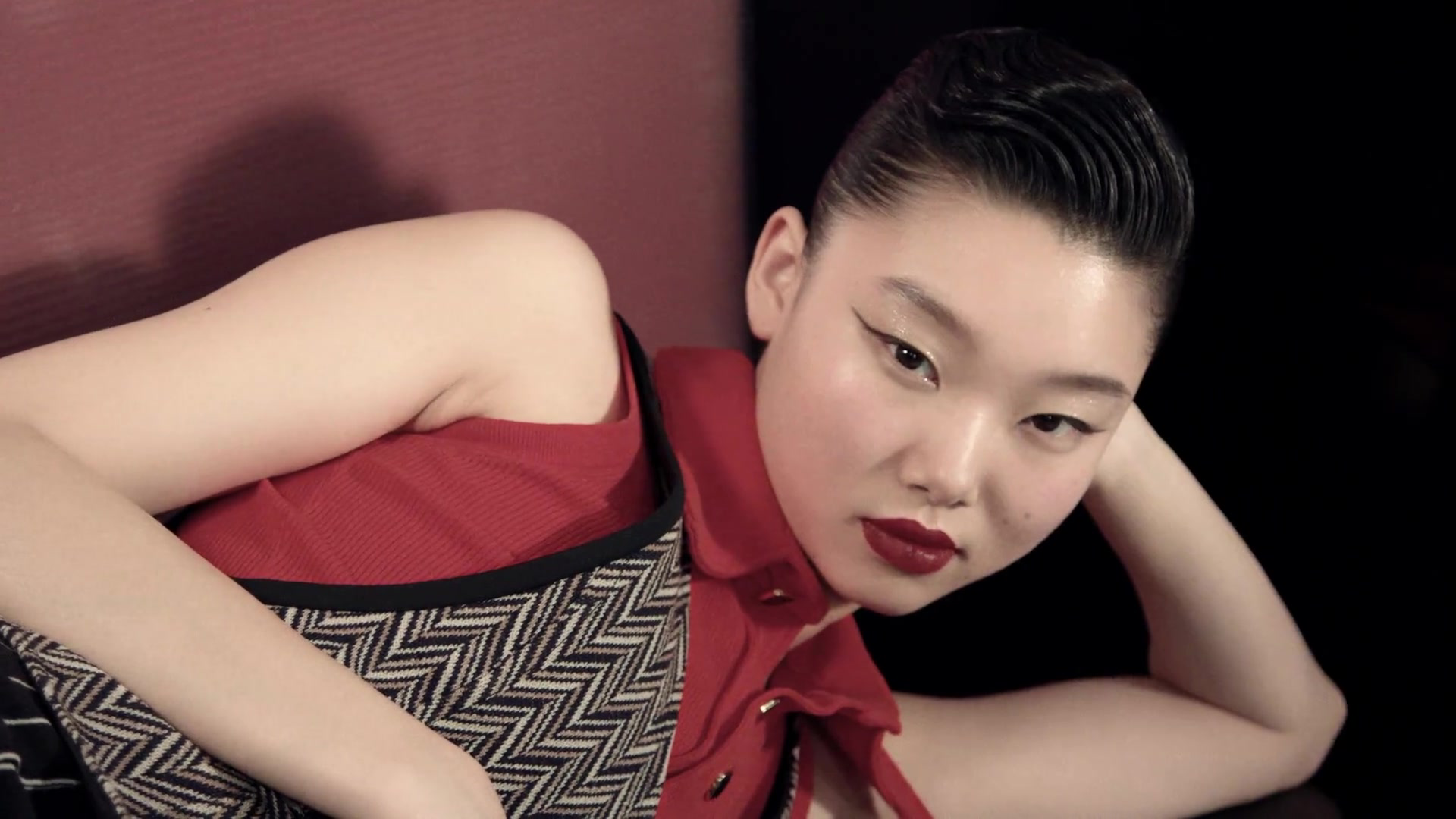Reclining person with slicked-back hair and red lipstick beside a red upholstered backrest